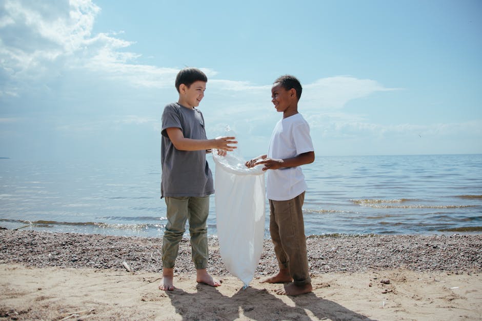 Two boys standing barefoot on a sandy beach near the water's edge, engaged in a playful activity involving a large transparent plastic bag filled with water. The boy on the left, wearing a gray T-shir